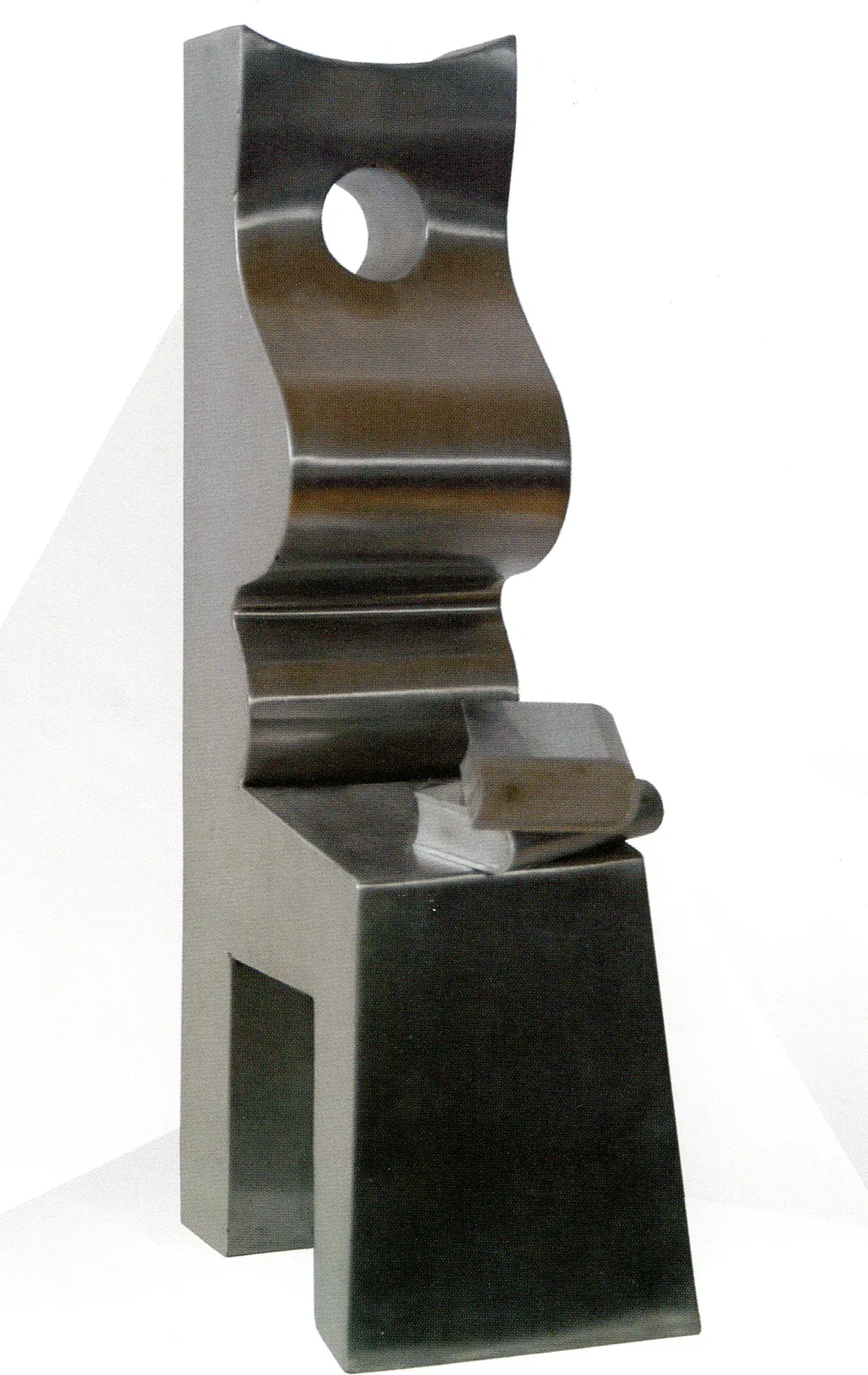 Scultura d’acciaio lucidato: una lastra verticale alta fa da schienale, davanti una superficie ondulata descrive un corpo seduto di profilo con un foro circolare al posto del capo; in basso il piano del grembo con due libri d’acciaio, e sotto un volume trapezoidale che fa da gambe