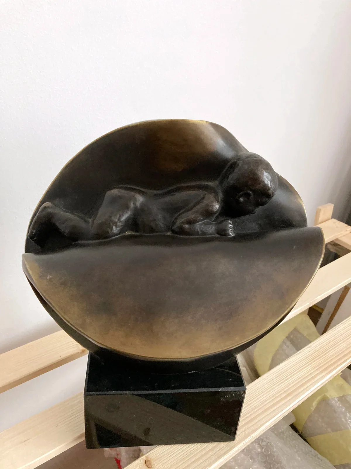 La stessa scultura vista da poco più in alto: il neonato si vede per intero, disteso sul piano chiaro del guscio aperto, con le gambe raccolte e un braccio steso in avanti