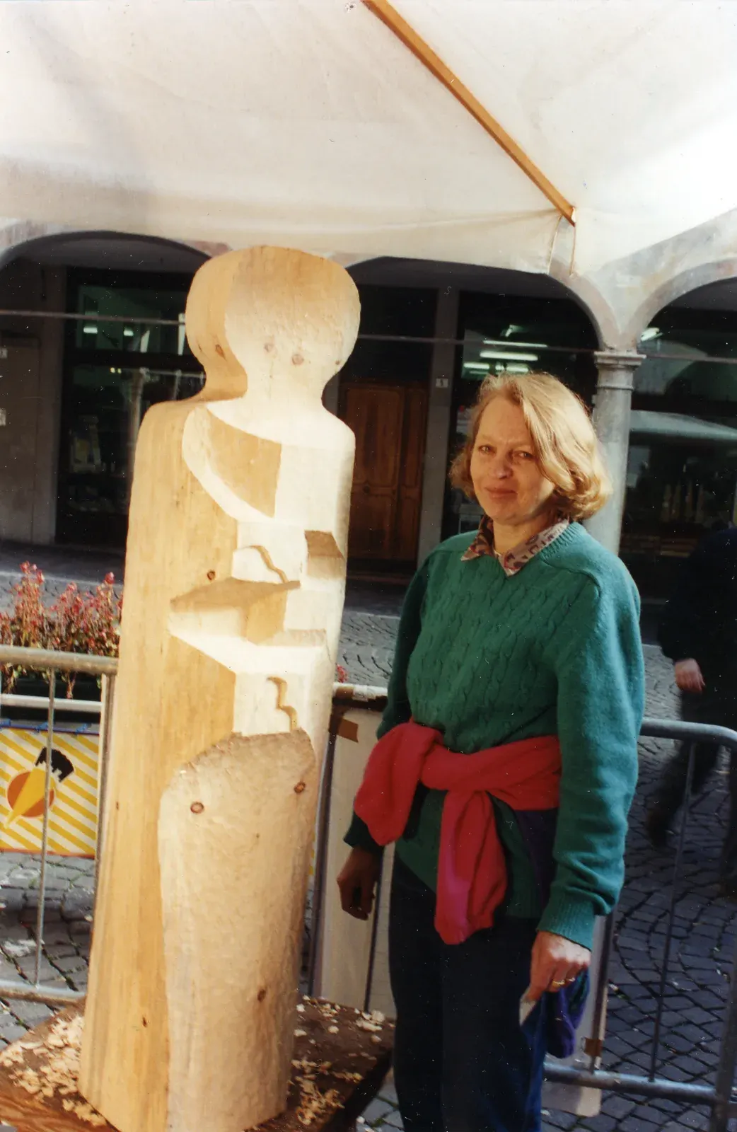 Dom — Antonina accanto alla scultura appena finita, sotto il telo bianco della postazione: dietro di lei i portici della piazza, ai piedi del tronco i trucioli. È la 9ª Ex Tempore di Belluno, novembre 1994.