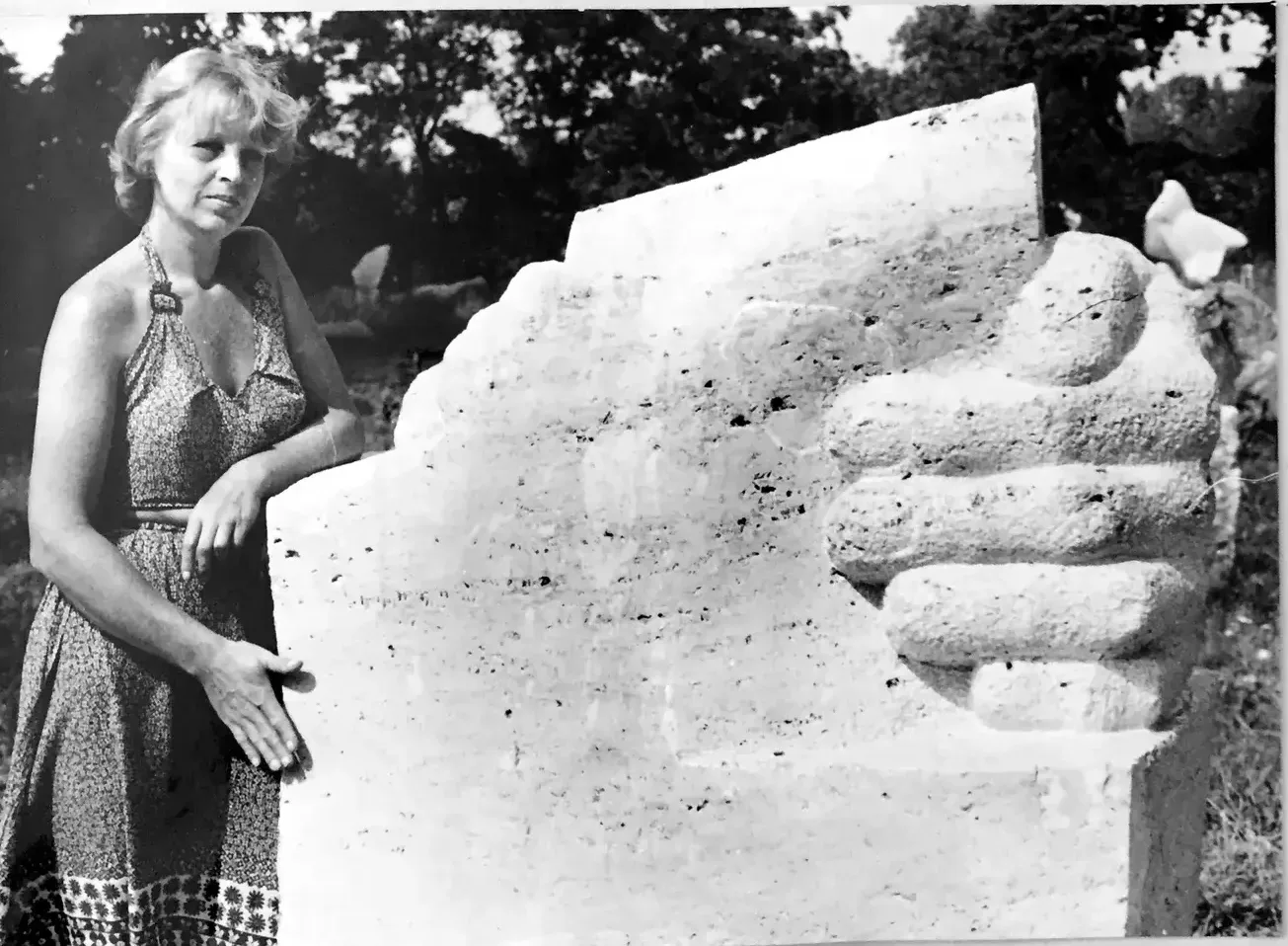 Antonina Wysocka-Jonczak in abito estivo accanto al blocco di travertino chiaro, la mano appoggiata alla pietra; la scultura, alta quanto lei, mostra sul fianco destro le quattro dita ripiegate