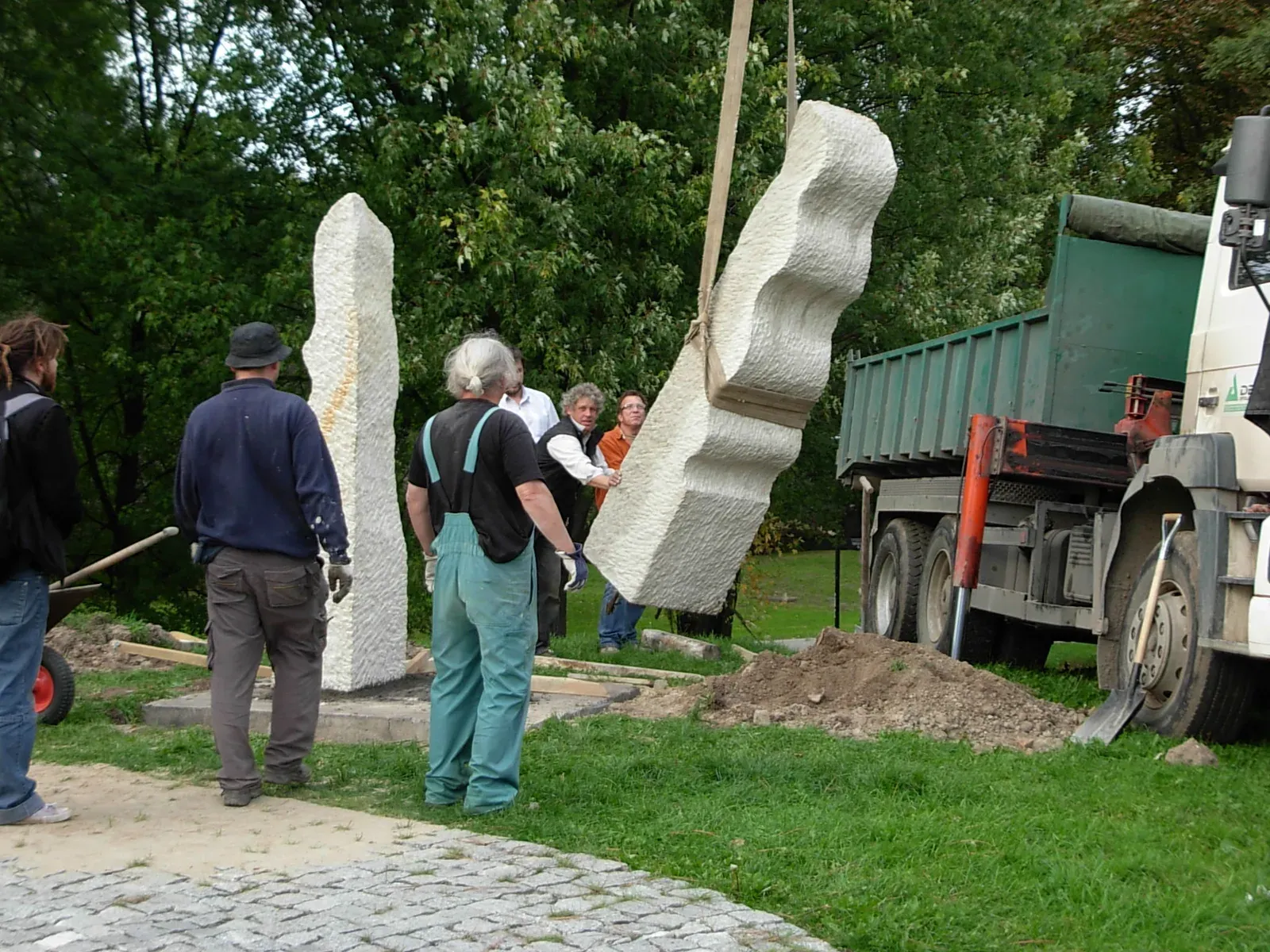 La posa in opera: una stele è già in piedi nel prato, l’altra pende dalle cinghie della gru di un camion mentre alcuni uomini la guidano verso la buca