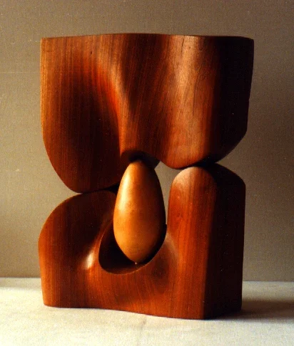 Piccola scultura di mogano lucidato: due masse sovrapposte, quella sopra che scende con una curva e quella sotto aperta da una nicchia, e nel varco un ovoide di legno chiaro levigato