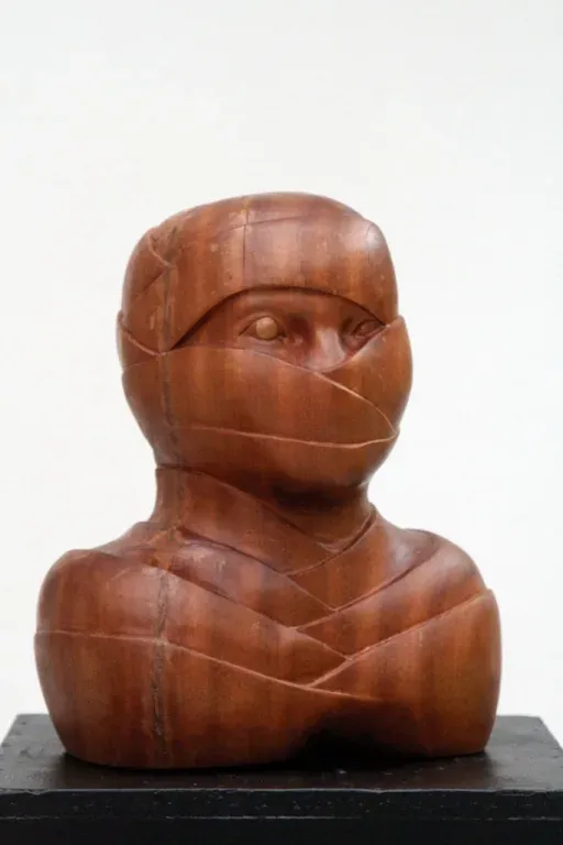 Subiektywny I: testa e busto di legno fasciati da bende scolpite che coprono naso e bocca e scendono sul petto; fra due bende resta scoperta una fessura all’altezza degli occhi, e in un occhio la pupilla è una sferetta sporgente di legno chiaro