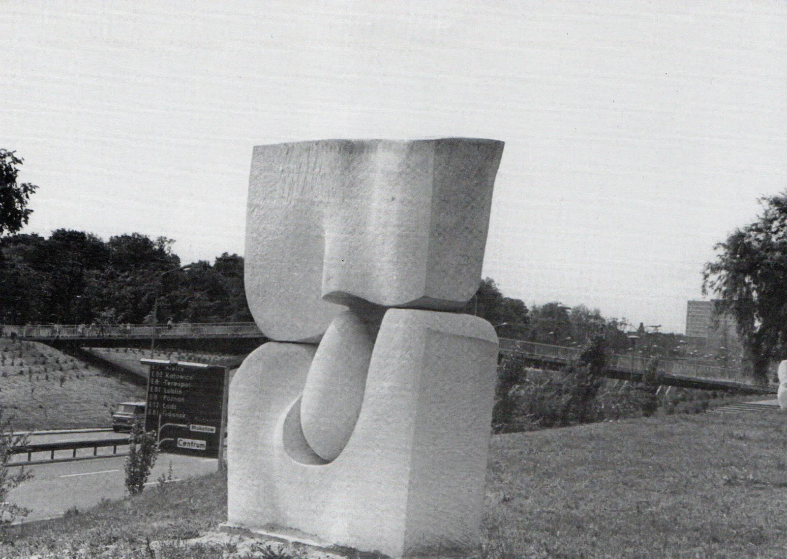 Grande scultura di pietra chiara su un prato accanto a una strada: un blocco squadrato diviso in due masse, quella superiore che scende con una curva e quella inferiore aperta da una nicchia, e nel varco fra le due un grande uovo di pietra levigato; sullo sfondo un viadotto e un cartello stradale