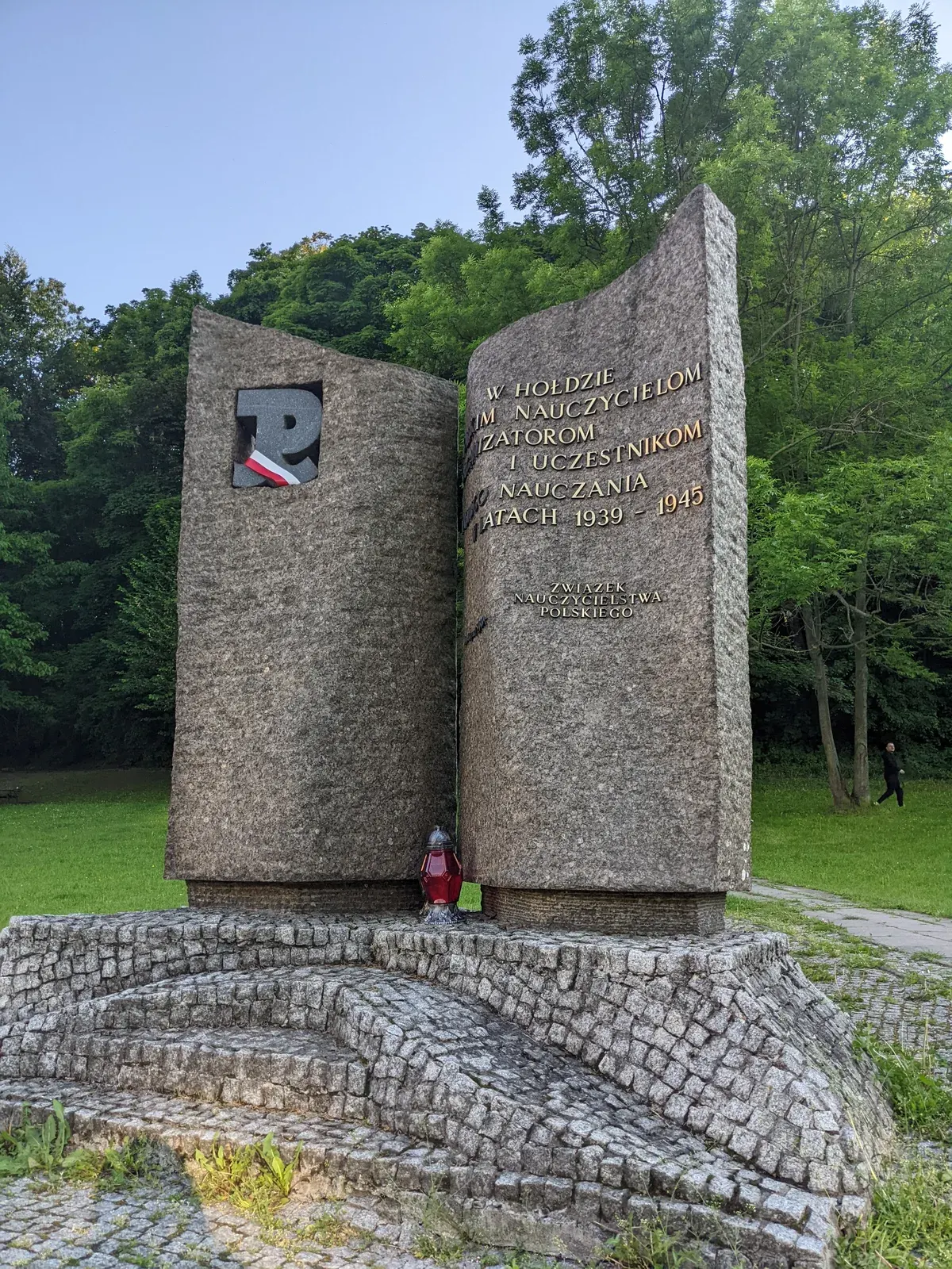 Pomnik Nauczycieli Tajnego Nauczania 1939–1945 — il monumento in assetto di cerimonia: un nastro bianco e rosso passato dentro la finestra, attraverso il segno della Polska Walcząca, e un lume rosso acceso ai piedi della fenditura, sul selciato a raggiera