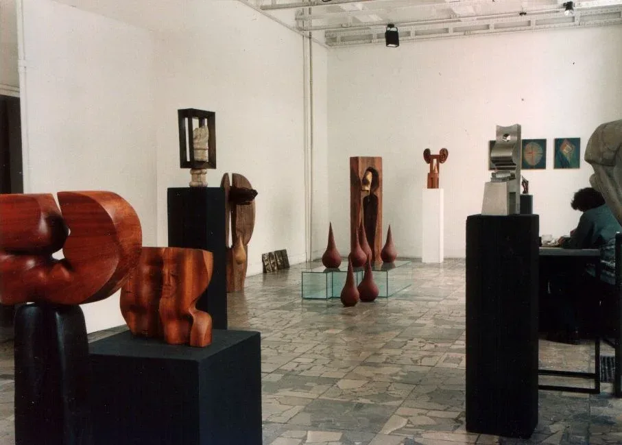Sala di galleria con pavimento di pietra: in primo piano, su plinti neri, due sculture di mogano — la seconda è Kolejne odbicia, con i suoi due volti; sul fondo altre sculture di legno e di pietra e una fila di forme a goccia su una teca di vetro