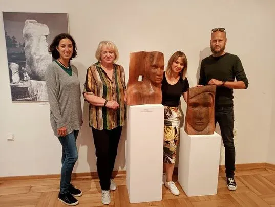 Quattro visitatori in posa accanto a due busti di legno sui plinti bianchi, davanti alla grande fotografia in bianco e nero appesa alla parete.