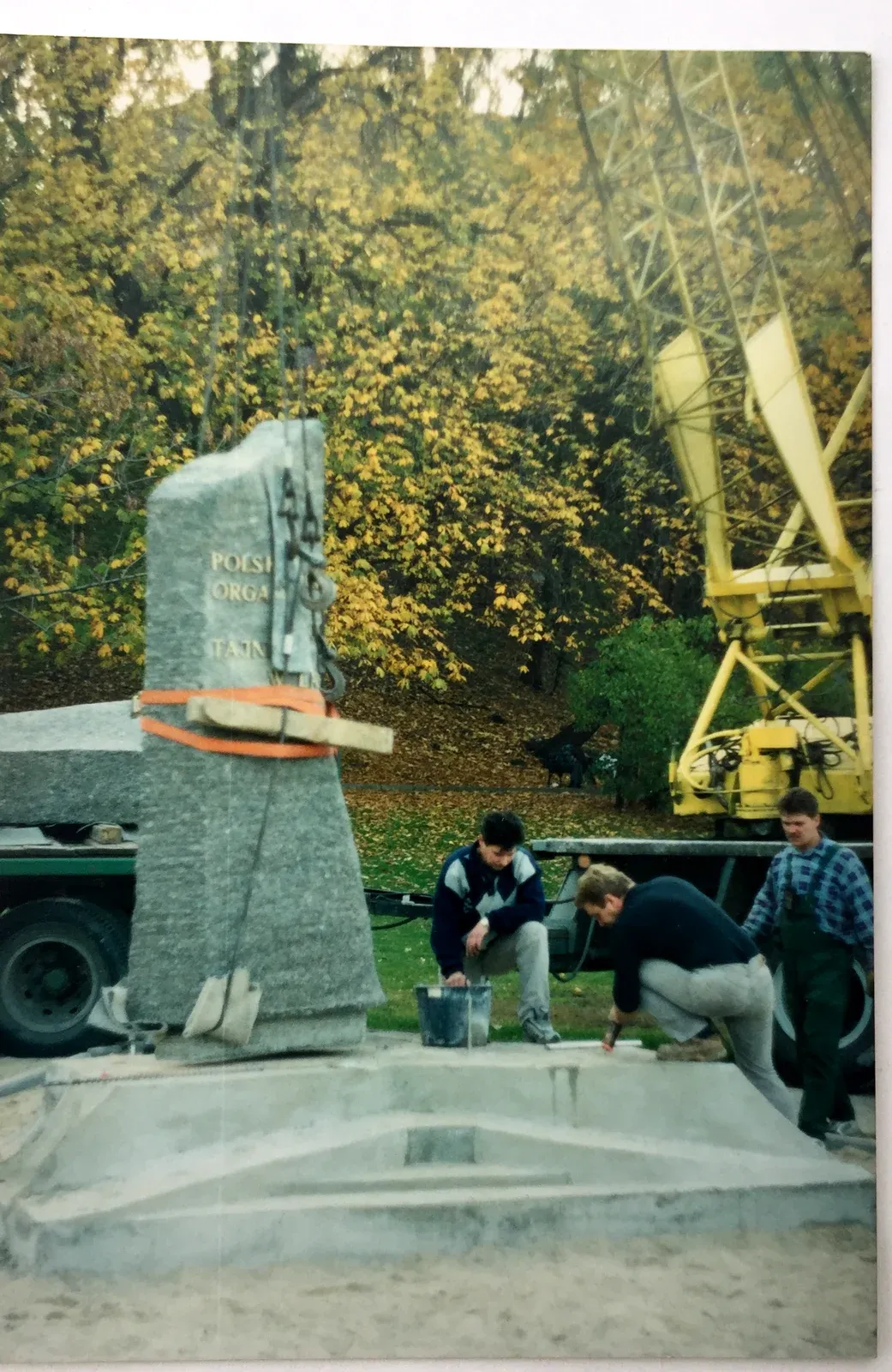 Tre operai al lavoro sulla base mentre la stele iscritta, ancora imbragata, tocca la sua posizione