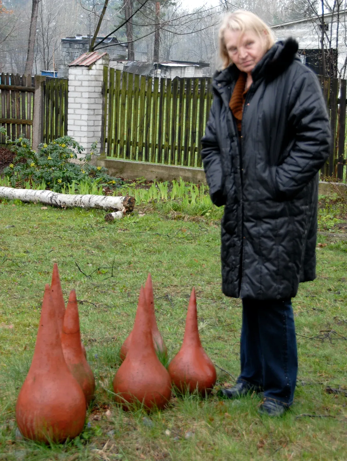 Antonina in cappotto scuro, in piedi in un giardino accanto a quattro gocce di ceramica invetriata di rosso vivo posate nell’erba